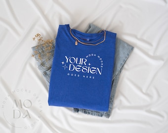 May include: A royal blue t-shirt with the text "YOUR DESIGN GOES HERE" in white, paired with light-wash denim jeans. A gold necklace and dried flowers are also present. The items are arranged on a white fabric background.