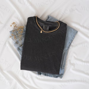 May include: A folded charcoal gray t-shirt with a gold necklace and a small pendant rests on top of a pair of light blue denim jeans. A small bundle of dried flowers is tucked into the jeans pocket. The items are arranged on a white fabric.