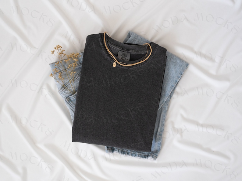 May include: A folded charcoal gray t-shirt with a gold necklace and a small pendant rests on top of a pair of light blue denim jeans. A small bundle of dried flowers is tucked into the jeans pocket. The items are arranged on a white fabric.