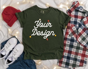May include: A green t-shirt with the text "Your Design" in white cursive lettering. The shirt is laid flat on a white surface with a red beanie, white sneakers, blue jeans, and a plaid shirt.