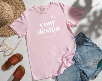 May include: A pink t-shirt with the text "Your design goes here" in white. The shirt is laid flat on a white surface with a straw hat, sunglasses, and denim shorts.