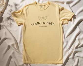 May include: A pale yellow t-shirt with a simple design of sunglasses and the text "YOUR DESIGN GOES HERE" on a white background.