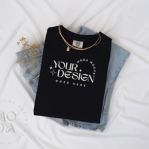 Op de afbeelding: Een zwart T-shirt met een witte grafische afbeelding waarop "YOUR DESIGN GOES HERE" staat en een gouden ketting. Het T-shirt is gevouwen en ligt op een blauwe spijkerbroek. De achtergrond is wit.