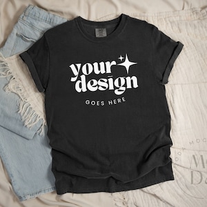 May include: Black t-shirt with a white graphic that says "your design goes here" with a star above the word "your". The shirt is laying on a white and blue fabric background.