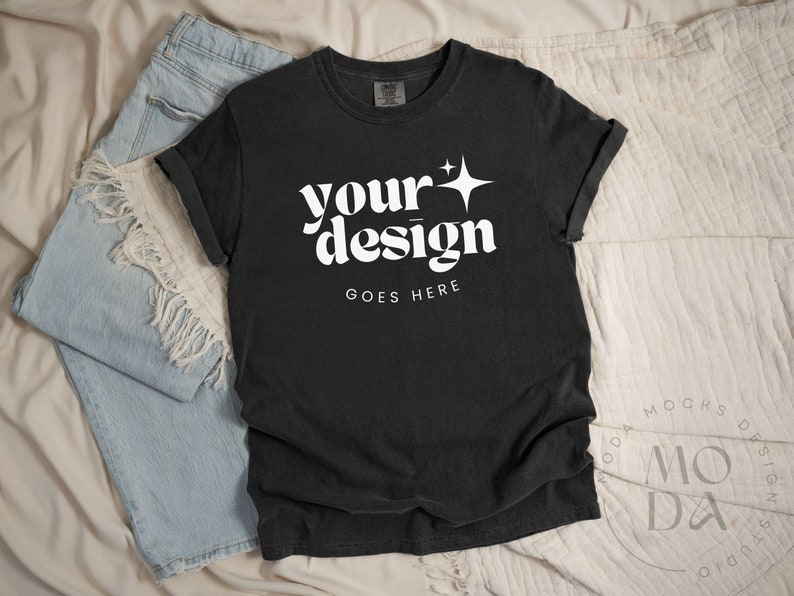 May include: Black t-shirt with a white graphic that says "your design goes here" with a star above the word "your". The shirt is laying on a white and blue fabric background.