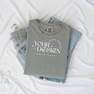 May include: A sage green t-shirt with the text "YOUR DESIGN GOES HERE" in a white circle with a star design. The t-shirt is laying on a white surface with a pair of blue jeans and dried flowers.