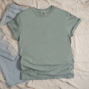 May include: A sage green t-shirt with rolled up sleeves, laying on a cream colored fabric background. The t-shirt is blank and has a tag on the bottom left side.