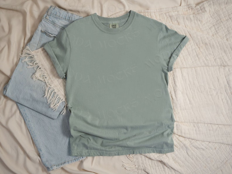 May include: A sage green t-shirt with rolled up sleeves, laying on a cream colored fabric background. The t-shirt is blank and has a tag on the bottom left side.