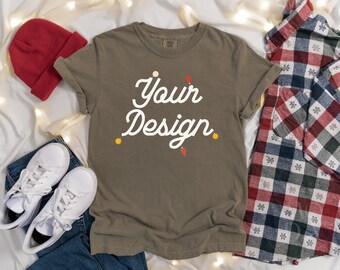 May include: A brown t-shirt with the text "Your Design" in white lettering. The shirt is laid flat on a white surface with a red beanie, white sneakers, blue jeans, and a plaid shirt.