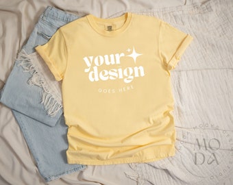 May include: A yellow t-shirt with the text "your design goes here" in white. The shirt is on a white and beige textured surface with blue jeans.