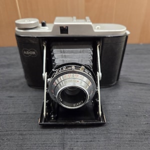 Puede incluir: Una cámara plegable ADOX vintage en negro y plata. La cámara tiene una parte superior y una carcasa de lente plateadas, un fuelle negro y un cuerpo negro. La lente está marcada con la marca ADOX y varios ajustes. La cámara está abierta y lista para usar.