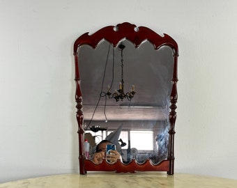 May include: A mahogany wood framed mirror with a bevelled glass mirror. The mirror has a scalloped top and a decorative carved design. The mirror is sitting on a white marble surface.