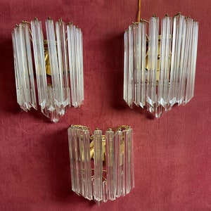 May include: Three clear glass wall sconces with gold metal accents, mounted on a burgundy wall.