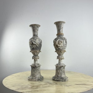 Könnte beinhalten: Zwei hohe, weiße Alabastervasen mit aufwendigen floralen Schnitzereien. Die Vasen stehen auf einem runden, weißen Marmortisch mit einem schwarzen Sockel.