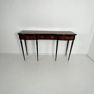 Könnte beinhalten: Eine Konsole aus dunklem Holz mit drei Schubladen und schlanken, sich verjüngenden Beinen. Der Tisch hat eine geschwungene Front und Messingbeschläge an den Schubladen. Die Beine haben Messingenden. Der Tisch steht vor einer weißen Wand und einem weißen Boden.