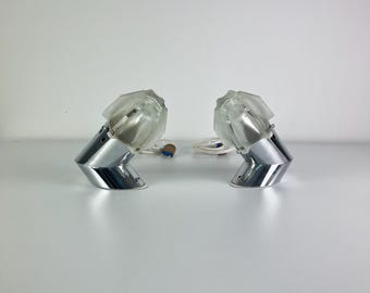 May include: Two silver-toned, geometric-shaped cufflinks with clear, faceted tops. The cufflinks are displayed on a white surface against a white background. The design features a modern, angular aesthetic.