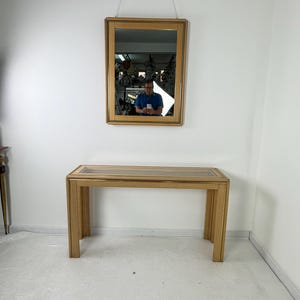 May include: A rectangular wooden framed mirror hangs above a matching wooden console table. The mirror is suspended by a chain. The console table has a glass top and a simple, modern design. The wood has a light, natural finish.