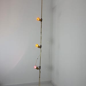 Può includere: Una lampada a sospensione vintage in ottone, con tre lampadine colorate. La lampada ha un design a spirale nella parte superiore e inferiore, con un lungo e sottile palo. Le lampadine emettono luce arancione e rosa, creando un'atmosfera calda.