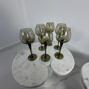 May include: Six smoky-grey wine glasses with long stems are arranged on a white marble table. The glasses have a rounded bowl shape. The table has a unique, organic design with a marble finish.