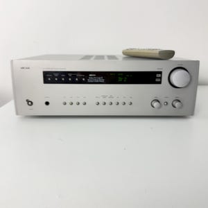 Pode incluir: Receptor estéreo prateado com controle remoto. O receptor possui uma tela e vários botões e botões. A marca "Arcam" é visível na frente. O controle remoto é bege.