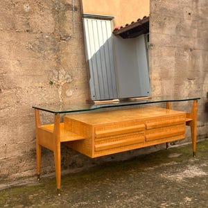 May include: A mid-century modern dresser with a glass top and four drawers. The dresser is made of light wood and has a simple, elegant design. It is sitting in front of a wall with a mirror hanging above it.