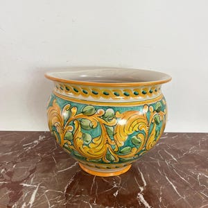 Peut inclure: Un pot de fleurs en céramique de forme large et arrondie. Le pot présente un motif floral jaune et vert vif sur un fond turquoise. Le bord et la base sont accentués d'orange. Une pièce décorative pour l'intérieur ou l'extérieur.