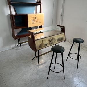 May include: Mid-century modern furniture set featuring a wall-mounted cabinet with a blue backing and a matching bar with a glass top. Includes two black stools with green seats. The furniture has a light wood finish with architectural illustrations.