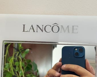 May include: A white mirror with a silver Lancôme logo at the top. A person is holding a dark blue smartphone in front of the mirror, reflecting a green plant.