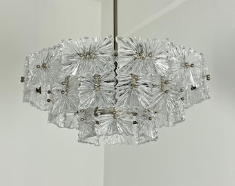 Puede incluir: Una lámpara de araña de cristal transparente con un diseño escalonado en forma de flor. La lámpara tiene múltiples capas de elementos de vidrio, cada uno con un patrón texturizado en forma de estrella. Los herrajes metálicos son de color dorado apagado.