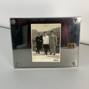 May include: A rectangular, silver-toned metal picture frame with a vintage black and white photograph of three men. The photo is of the men standing outside with a mountain backdrop. The frame has four small, square magnets.