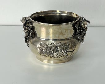 Puede incluir: Enfriador de vino plateado con detalles florales y racimos de uvas. El enfriador tiene dos asas con diseños de racimos de uvas. El cuerpo del enfriador presenta un patrón floral detallado. El enfriador está hecho de metal brillante y reflectante.