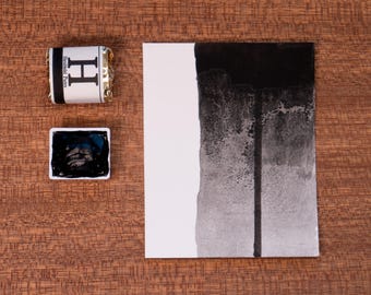 May include: A black watercolour paint pan with a white label that says "Daniel Smith -  H -  Ivory Black" and a black watercolour wash on a white paper.