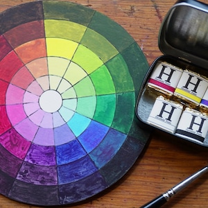 May include: A colour wheel with a variety of colours, including red, orange, yellow, green, blue, indigo, and violet. There are five small paint pans with the letter "H" on them, and a paintbrush.