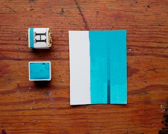 Può includere: Un piccolo vassoio quadrato di acquerello blu turchese con la lettera "H" sull'etichetta. Un cartoncino bianco con una striscia blu turchese dipinta su di esso.