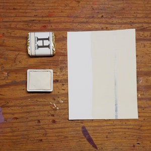 May include: A white watercolour pan with a gold trim sits on a wooden surface next to a closed watercolour pan with a gold trim and the letter "H" printed on the front. A white sheet of paper with a grey line down the centre is on the right side of the image.