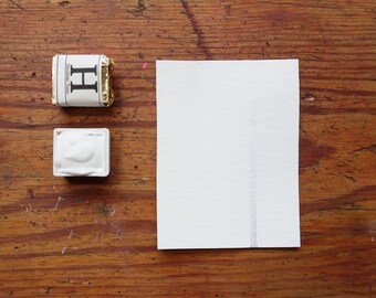 May include: Two watercolour paint pans on a wooden surface. The pan on the left is gold with the letter "H" printed on it. The pan on the right is white with a white paint inside. A white sheet of paper is to the right of the pans.