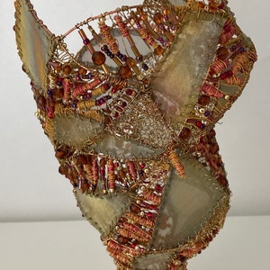 May include: A decorative sculpture made of wire, fabric, and beads. The sculpture is in the shape of a vase with a wide mouth and a narrow base. The fabric is a light brown color and has a textured surface. The beads are a variety of colors, including red, orange, and brown. The sculpture is a unique and eye-catching piece of art.