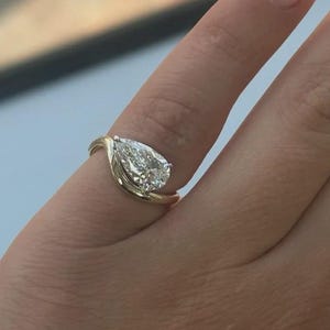 May include: A gold ring featuring a pear-shaped diamond. The diamond is the focal point, set in a swirling gold band. The ring is shown on a finger, highlighting its design and sparkle.