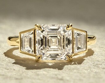 Può includere: Un anello d'oro con un grande diamante centrale a taglio quadrato, affiancato da due diamanti rettangolari più piccoli. La montatura è in oro lucido. I diamanti brillano. Un gioiello elegante.