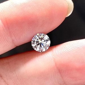 May include: A round, clear diamond gemstone with a brilliant cut. The diamond is set in a ring and is held by a hand.
