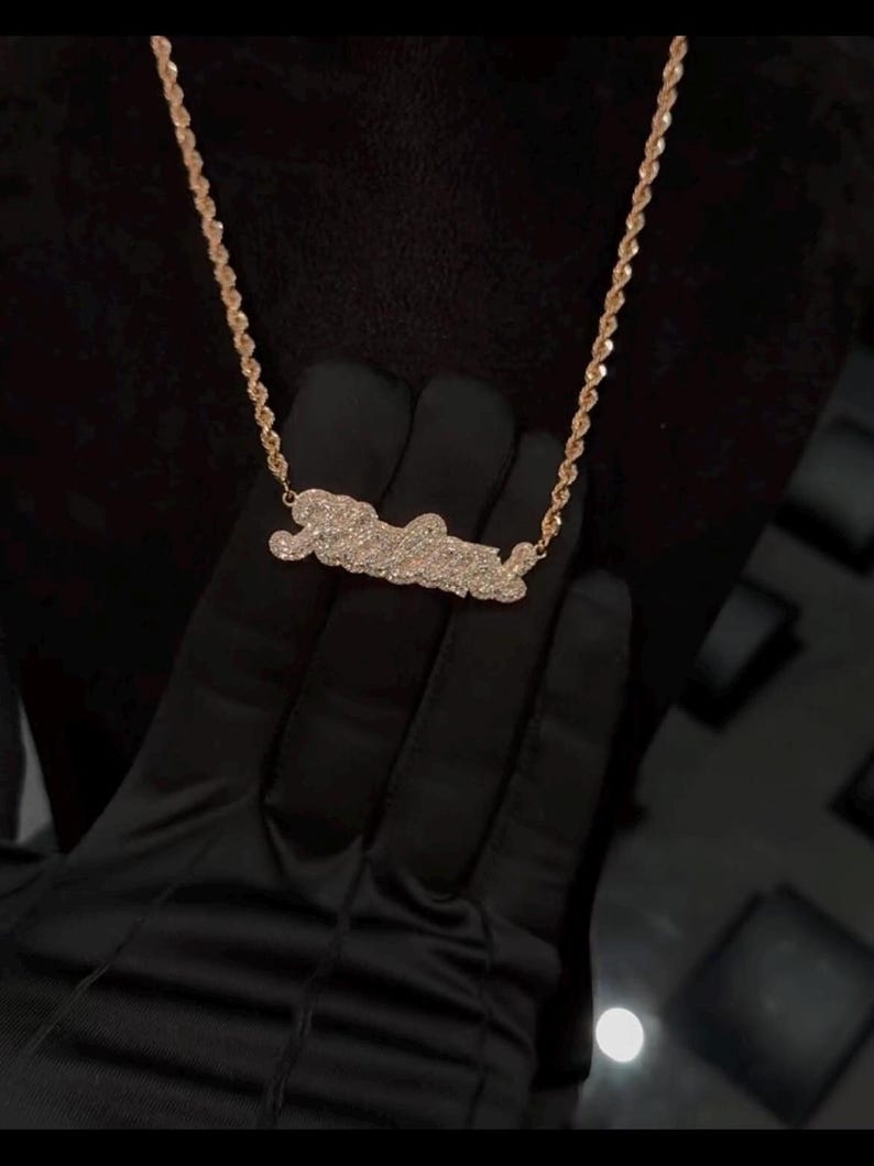 May include: A gold chain necklace with a diamond-encrusted pendant in the shape of a word. The pendant is a popular style for personalized jewelry.