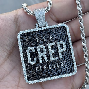 May include: Silver-toned pendant necklace with a square pendant. The pendant features the words "THE CREP CLEANER" in white, outlined with clear stones, set against a black background. The pendant is framed with clear stones and hangs from a silver rope chain.