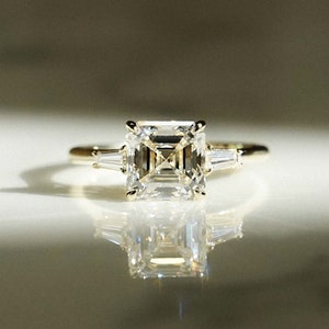 May include: A gold ring with a large, square-shaped, clear emerald-cut diamond and two smaller, baguette-cut diamonds on either side.