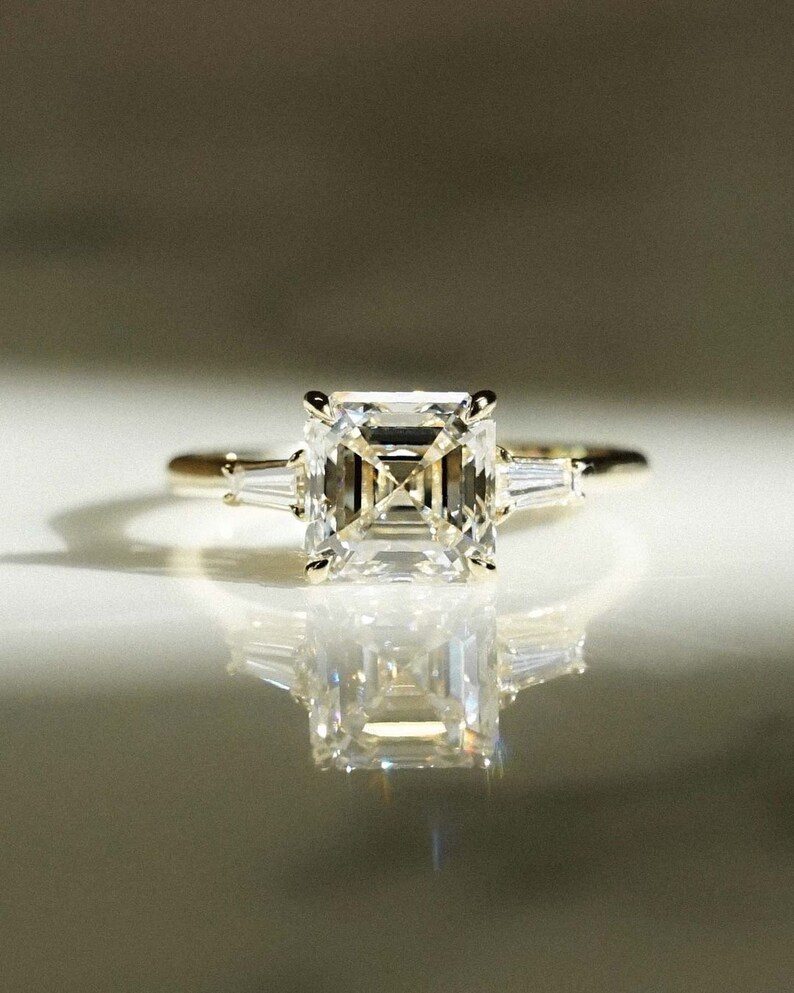 May include: A gold ring with a large, square-shaped, clear emerald-cut diamond and two smaller, baguette-cut diamonds on either side.