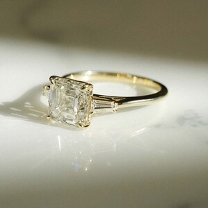 May include: A gold engagement ring with a square-shaped diamond and two smaller baguette diamonds on the band.