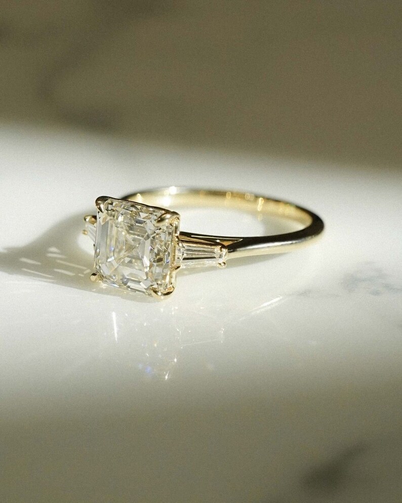 May include: A gold engagement ring with a square-shaped diamond and two smaller baguette diamonds on the band.
