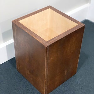 May include: A square wooden wastebasket with a light brown interior. The wastebasket is made of dark brown wood and has a smooth finish.