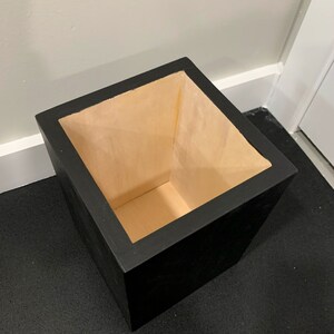 May include: A black square planter with a light brown wooden interior. The planter is empty and sitting on a black surface.