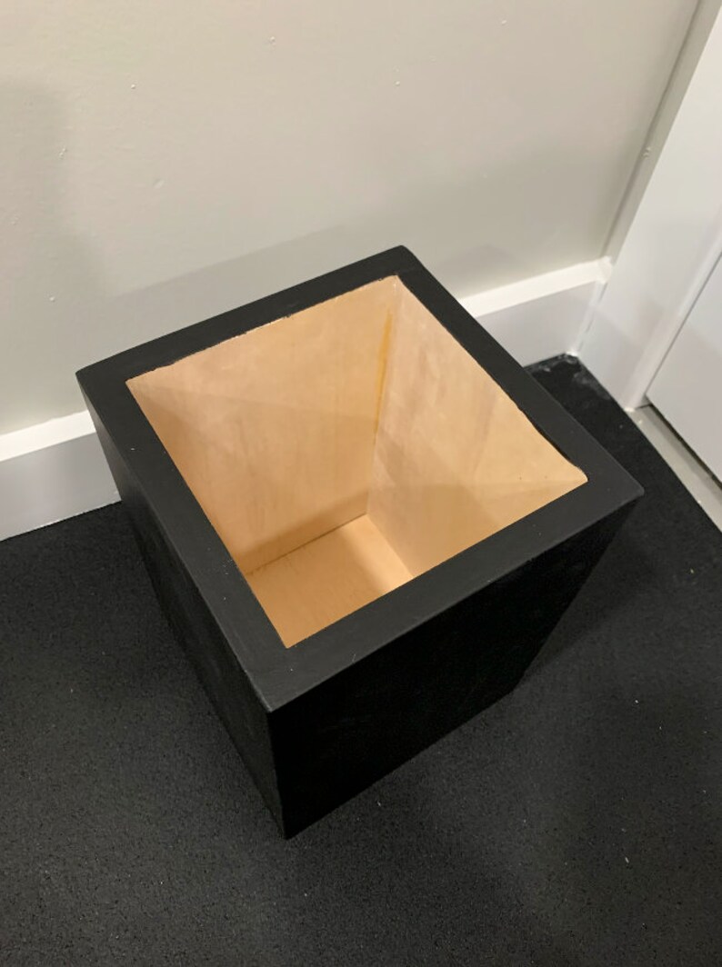 May include: A black square planter with a light brown wooden interior. The planter is empty and sitting on a black surface.