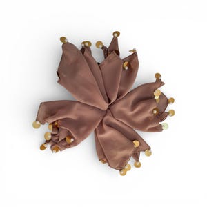 May include: A brown, flower-shaped accessory with a central point and multiple petal-like sections. Each petal is adorned with gold sequins and small, round, white beads. The accessory is set against a white background.